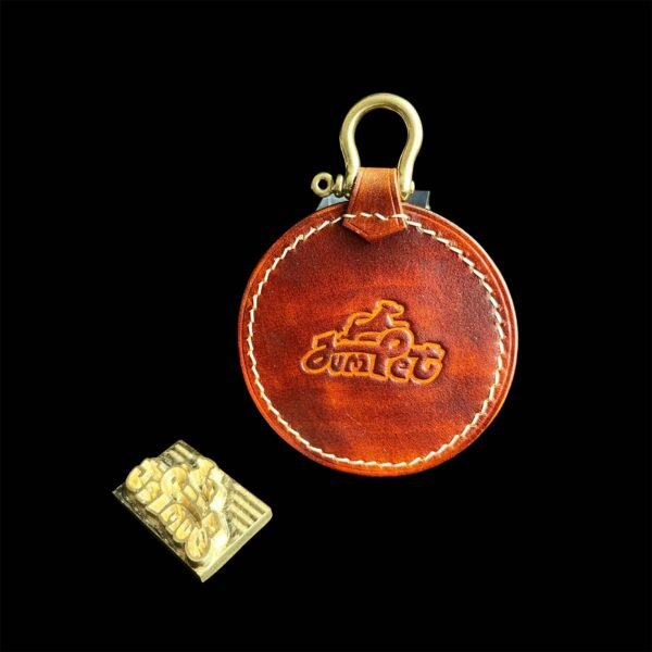 Against a black background, a custom pet portrait keychain made of round, brown leather with embossed writing and a brass clip sits beside a small gold-toned metal plaque showcasing two vehicles. This personalized leather piece makes for an exceptional Christmas present for pet lovers.