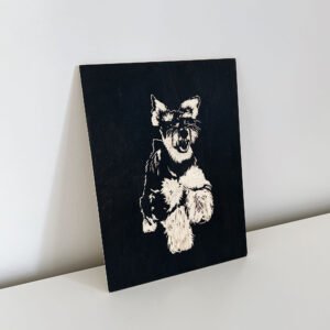 A Custom Wooden Engraved Pet Portrait, showcasing a leaping dog, possibly a terrier, with striking dark and light contrasts, is leaning against a plain white wall on a white surface. This handmade, eco-friendly memorial gift captures the playful energy of the pet beautifully.